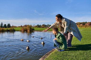 cheerful african american woman in outerwear pointing at ducks in pond near son, autumnal nature clipart