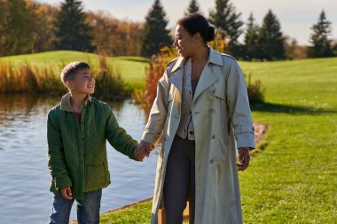happy mother and son holding hands and walking near lake, african american family, scenic view clipart