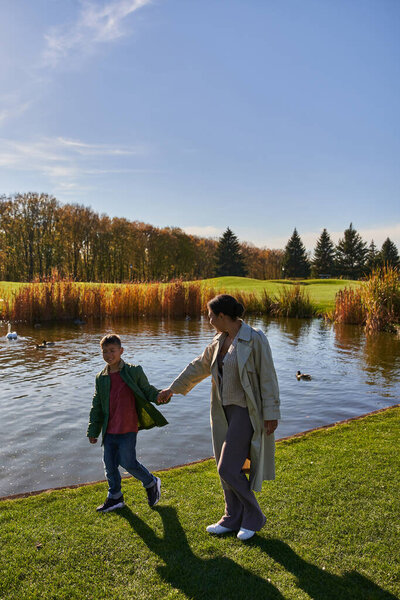 family bonding, autumnal nature, cheerful african american mother walking with son along pond