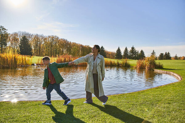 family bonding, happy african american woman following son, walking along pond, hold hands, autumn