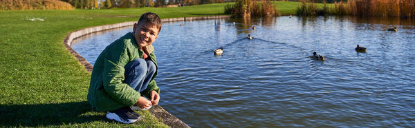 happy african american boy in outerwear sitting near pond with ducks, nature and kid, banner