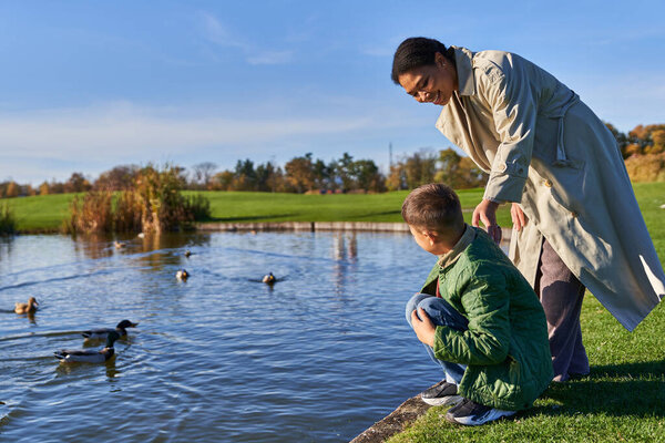 Осень, веселая африканская американка в верхней одежде стоит рядом с сыном рядом с прудом с утками