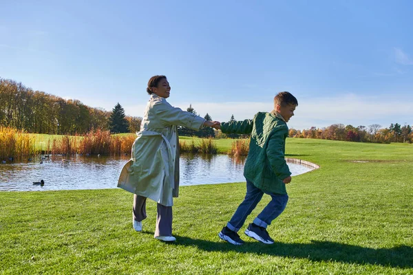 stock image playful african american mother and son running on grass near pond, modern parenting, fall fashion
