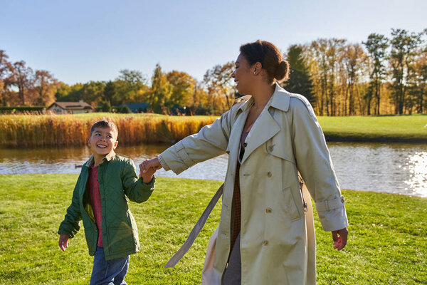 happy african american mother holding hands with son, walking near pond, modern parenting, fall