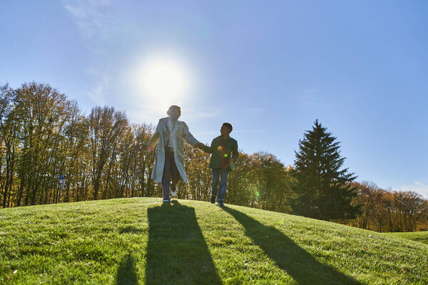 Веселая африканская мать и ребенок держатся за руки, бегают по зеленому холму, осенние наряды