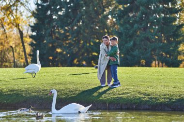 happy african american mother and son looking at swans in lake, nature, autumnal fashion, outerwear clipart