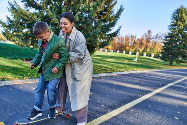 happy mother and son in autumn park, african american woman teaching boy how to ride penny board clipart