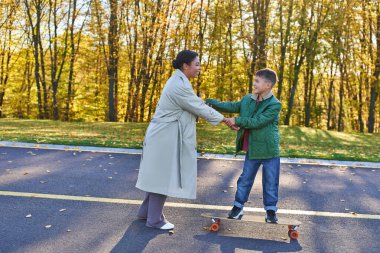 mother and son in park, joyful african american woman holding hands with boy on penny board, autumn clipart