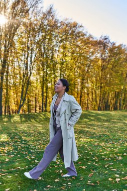 happy african american woman in trench coat walking on grass with fallen leaves, autumn, fashion clipart