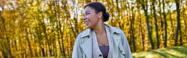 cheerful african american woman in trench coat walking in autumn park, fashion outfit, banner clipart