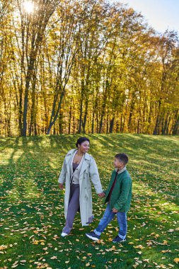 cheerful african american woman holding hands with son, walking on grass with golden leaves, autumn clipart