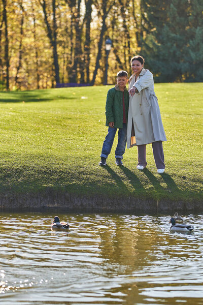 happy african american mother and son standing near lake, woman pointing at duck, nature, autumn
