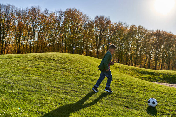 happy african american boy in autumnal clothes running on green grass, playing football, soccer ball