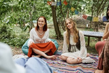 smiling and trendy women in boho styled outfits sitting on carpet outdoors in retreat center clipart