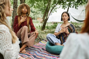 Çok ırklı kadınlar şık boho kıyafetleriyle inziva merkezindeki çayır üzerinde meditasyon yapıyorlar.