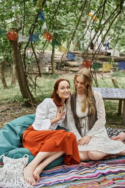 positive redhead woman in boho styled clothes sitting near friend on meadow in retreat center clipart