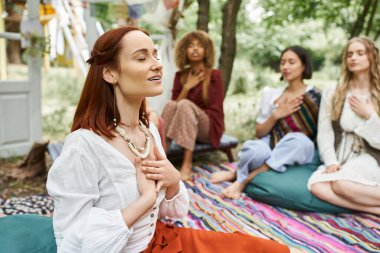 Neşeli kızıl saçlı kadın, dışarıda, kapalı alanda bulanık etnik arkadaşların yanında meditasyon yapıyor.