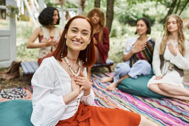 smiling and trendy redhead woman sitting near blurred multiethnic friends in retreat center clipart