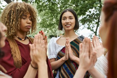 Boho giysileri içinde pozitif genç bir kadın inziva merkezindeki çok ırklı arkadaşlarının yanında meditasyon yapıyor.
