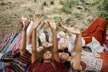 smiling interracial women raising hands while lying on blanket outdoors in retreat center clipart