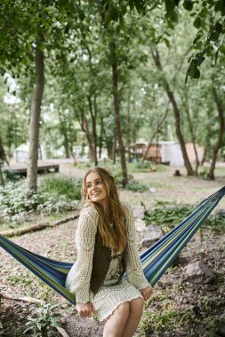 Gülümseyen, boho elbiseli genç sarışın kadın hamakta dinlenme merkezinde oturuyor.