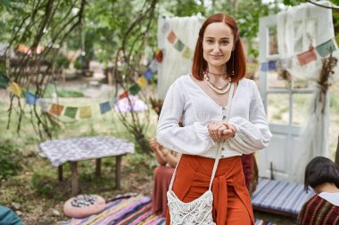 Boho kıyafetli pozitif kızıl saçlı bir kadın inziva merkezinde kameraya bakıyor.