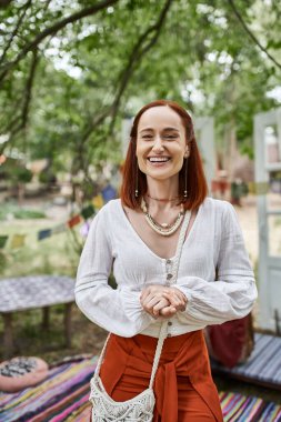 Boho kıyafetli heyecanlı kızıl saçlı kadının portresi. Dışarıda, dinlenme merkezindeki kameraya bakıyor.