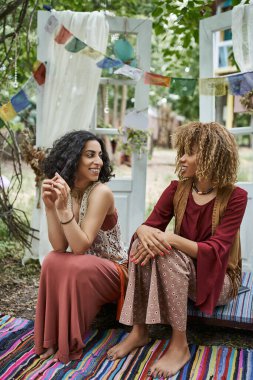 Boho kıyafetleri içinde gülümseyen çok ırklı kadınlar dışarıda dinlenme merkezinde vakit geçirirken konuşuyorlar.