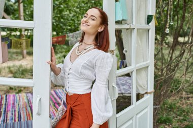 positive and trendy redhead woman in boho styled outfit standing outdoors in retreat center clipart