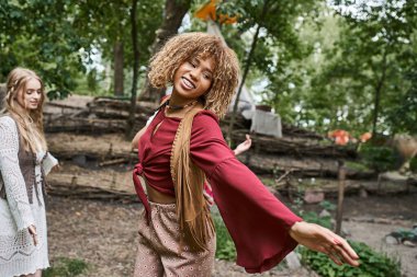 Genç ve gülümseyen Afrikalı Amerikalı kadın Boho kıyafetiyle inziva merkezinde çayırda dans ediyor.