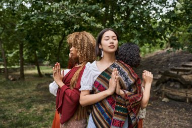 Genç ırklar arası kadınlar dua ederken, dışarıda dinlenme merkezinde dikilirken el hareketi yapıyorlar.