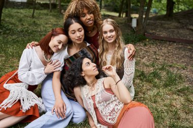 positive multiethnic women in boho outfits hugging while sitting on meadow in retreat center clipart