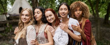 positive multiethnic women in boho outfits hugging outdoors in retreat center, banner clipart