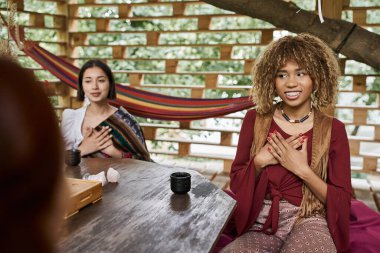 Gülümseyen Afrikalı Amerikalı kadın kız arkadaşlarının yanında meditasyon yapıyor ve çay fincanları ahşap masada.