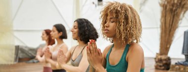 Genç Afrikalı Amerikalı kadın dua eden elleri ile çok ırklı kız arkadaşlarının yanında meditasyon yapıyor, afiş.
