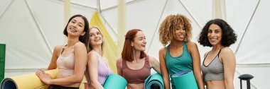 Kadınlar geri çekilme konsepti, spor giyimli çok ırklı kız arkadaşlar yoga minderleri ve afişle ayakta duruyorlar.