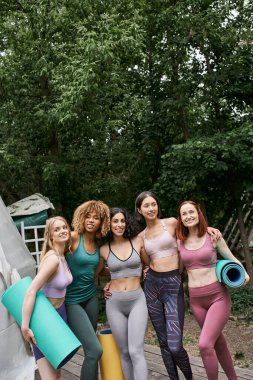 happy multiethnic women in sportswear standing with yoga mats in outdoor retreat center clipart
