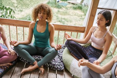 young african american woman meditating in lotus pose near girlfriends in cozy patio, tranquility clipart