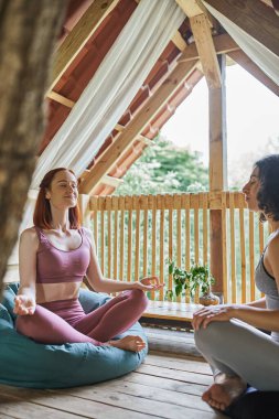 smiling woman with closed eyes sitting in lotus pose near multiracial girlfriend in cozy patio clipart