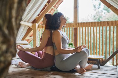 Spor kıyafetleri içinde, Lotus pozunda meditasyon yapan, sırt sırta oturan çok ırklı kız arkadaşlar.