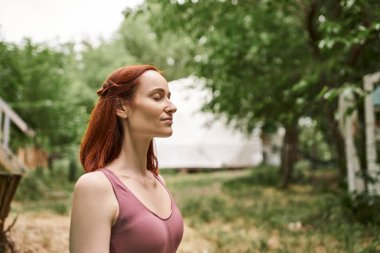 Mutlu kızıl saçlı kadın, kapalı gözlerle meditasyon yapıyor açık hava inziva merkezinde