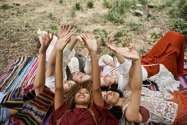 smiling interracial women raising hands while lying on blanket outdoors in retreat center