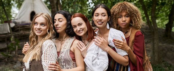 positive multiethnic women in boho outfits hugging outdoors in retreat center, banner