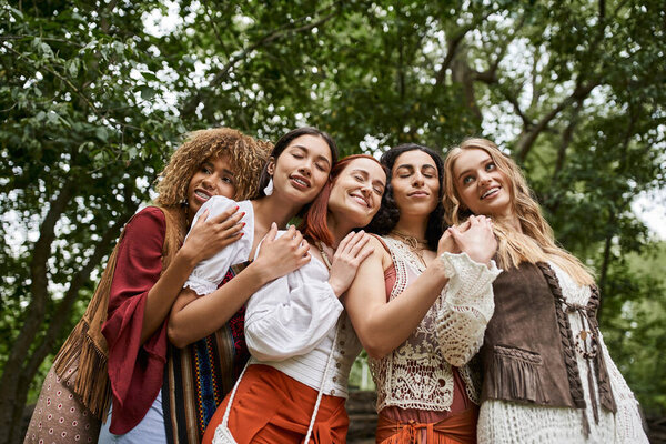low angle view of smiling multiethnic women in boho outfits hugging in retreat center