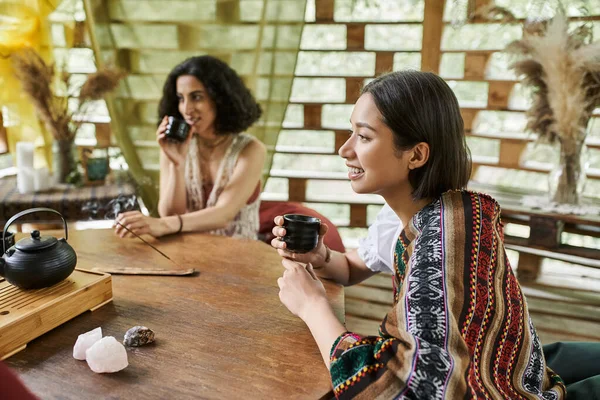 stock image cheerful young woman with tea cup near multiracial girlfriend at wooden table in retreat center