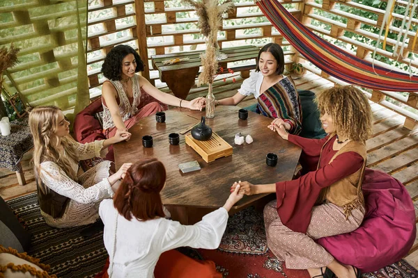 stock image smiling multiracial girlfriends holding hands and meditating at wooden table with tea cups