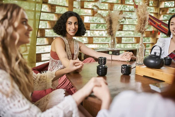 stock image happy multiracial woman holding hands and meditating with girlfriends near tea on wooden table