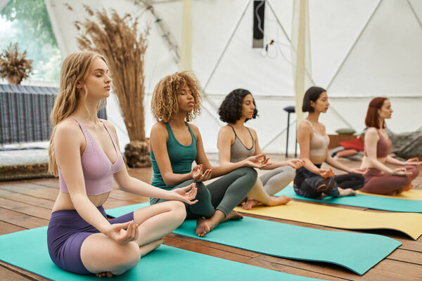 multiethnic girlfriends in sportswear meditating in lotus pose with closed eyes in retreat center