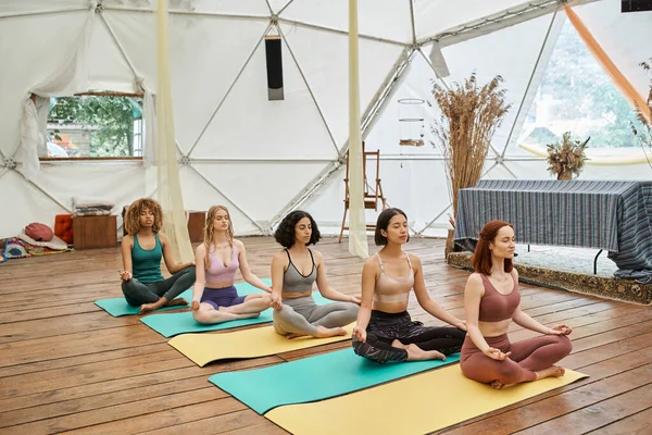 retreat center, multiethnic women on yoga mats, with closed eyes, practicing yoga in lotus pose