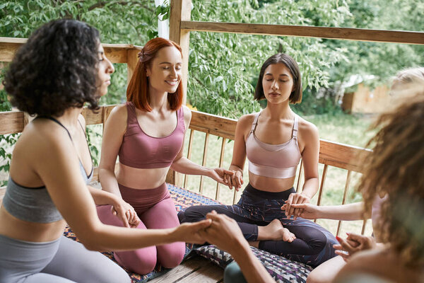 multiracial girlfriends in sportswear holding hands, sitting, meditating with closed eyes in patio
